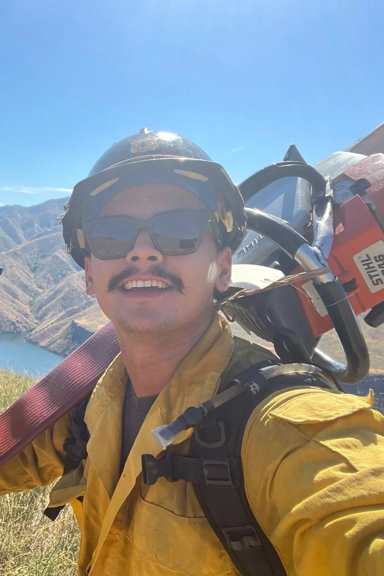 Per Gutierrez in wildland firefighting gear, helmet and yellow Nomex, carrying a chainsaw, on a hillside above a river canyon.